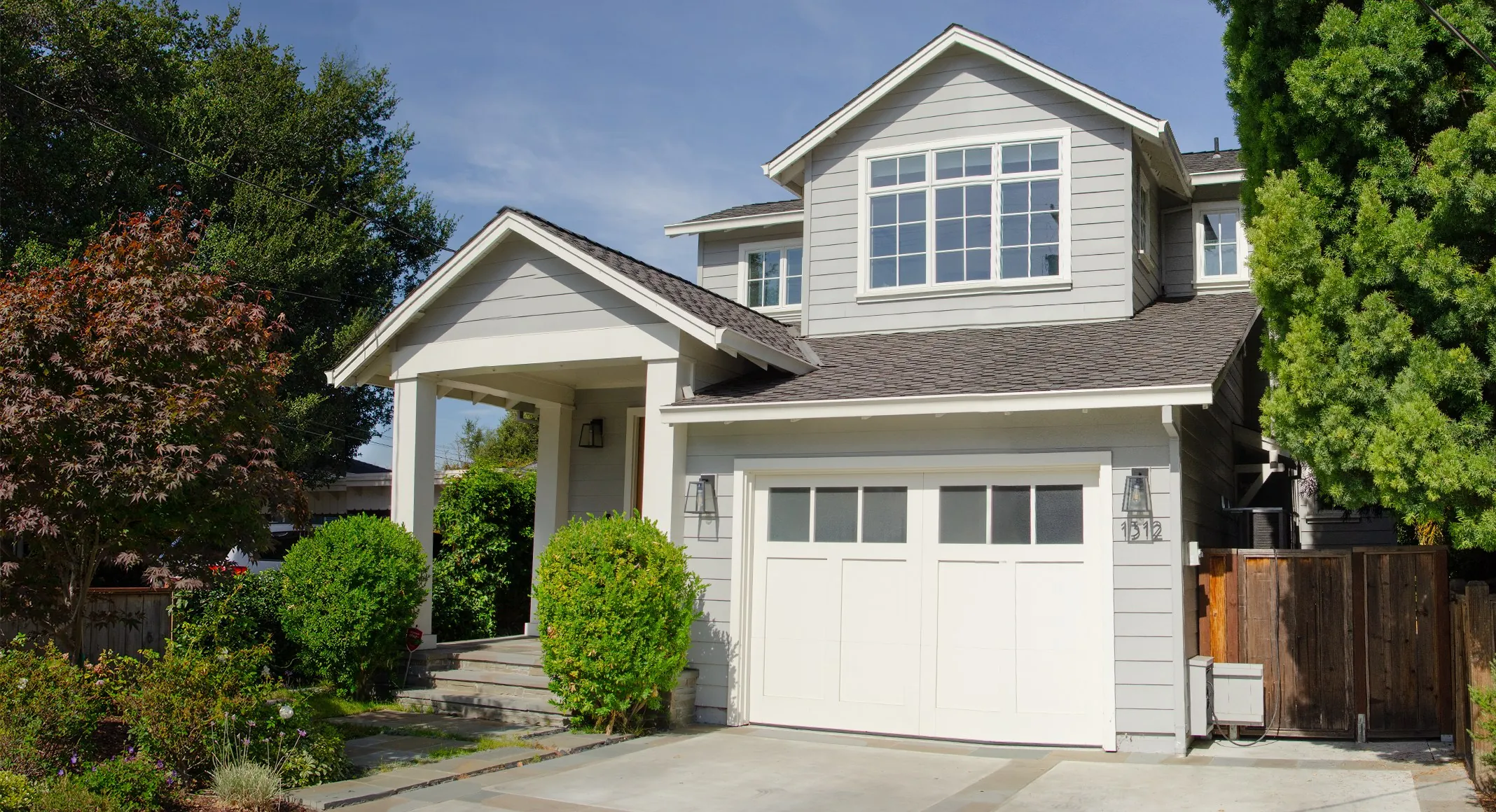 Wide front view of a San Carlos residence with gray siding, white trim, a covered entry porch, a gabled upper story, and a white garage door.